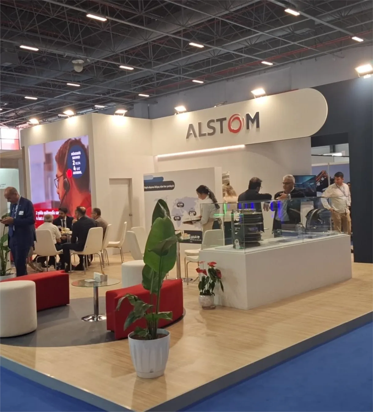 Alstom standı: kavisli beyaz alınlık, beyaz teşhir vitrinleri, LED ekran ve ahşap zemin üzerinde kırmızı-beyaz oturma grubu